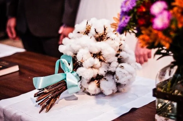 Buquê de flores de algodão com fita azul-petróleo sobre a mesa de um casamento, ao lado de um arranjo de flores