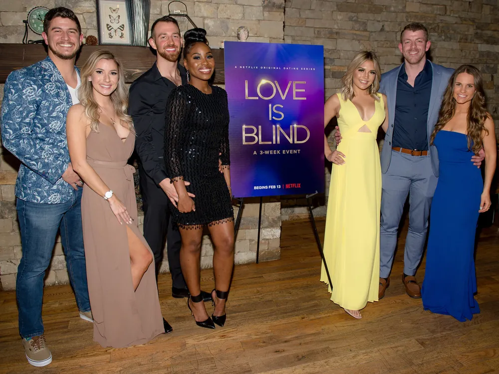 Grupo de participantes do Love Is Blind em trajes formais posando com um pôster do reality