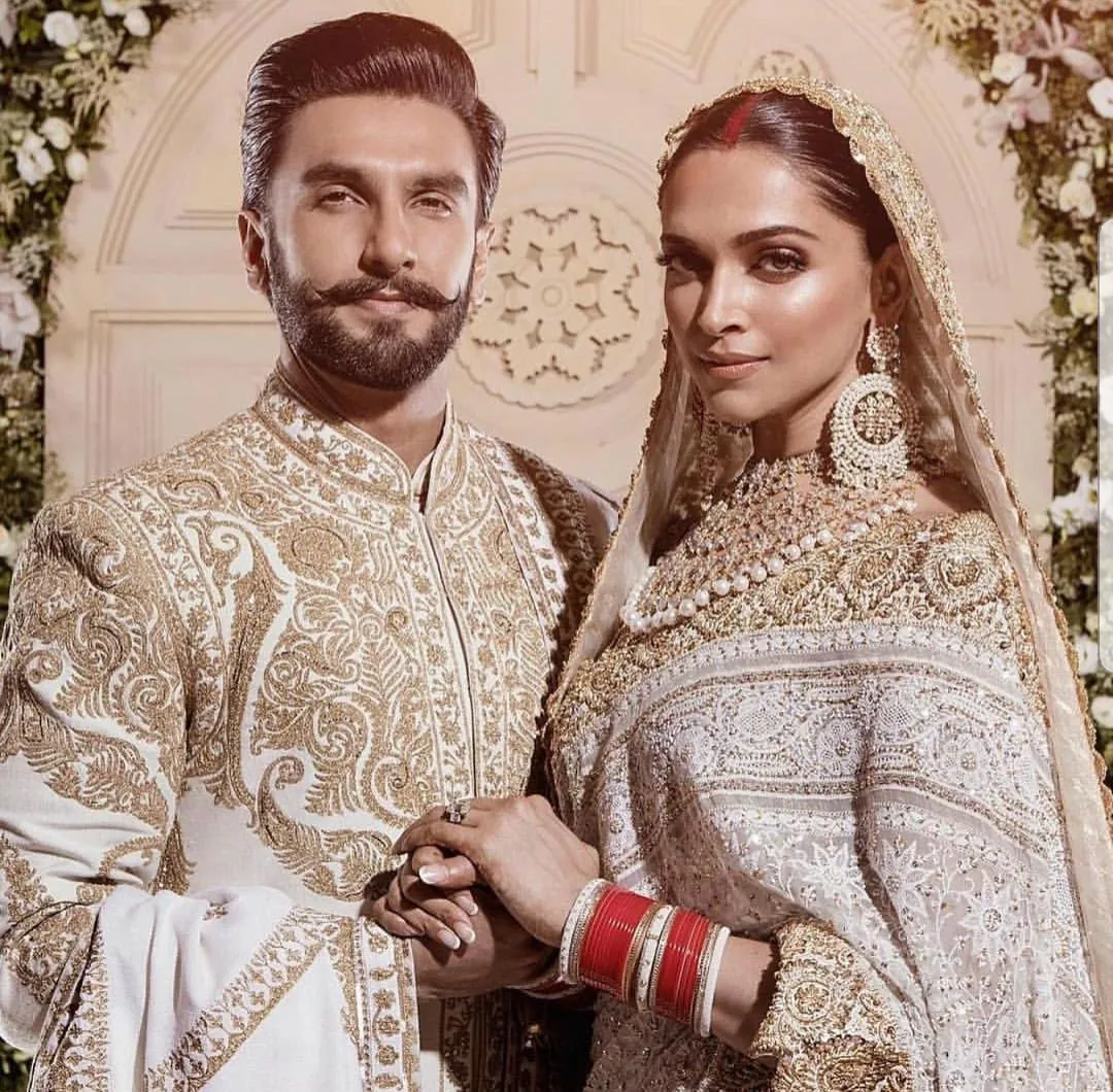 Casal em trajes tradicionais indianos bordados, de mãos dadas, com a noiva usando joias e pulseiras vermelhas