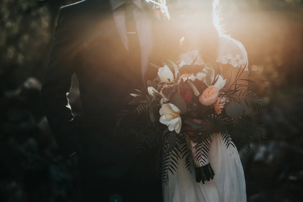 Casal de noivos de mãos dadas ao ar livre, com o sol forte atrás deles; a noiva segura um buquê de rosas e flores brancas com folhagens verdes